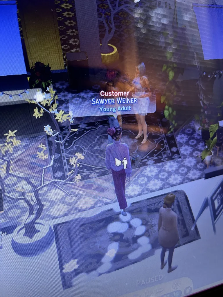 Photo of a Sims 4 gameplay screen showing a customer named “Sawyer Weiner” labeled as a Young Adult, standing inside a decorated venue while the game is paused.